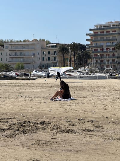 The image depicts a beach in Anzio, Italy, with the shoreline oriented vertically in the frame. In the foreground, a woman with dark hair is seated on a striped towel, appearing to be using a mobile device. Behind her is a sandy beach with a coarser texture. To the left of the beach are buildings and palm trees, and a row of sailboats and other small boats are parked on the sand near the buildings. The sky is clear and blue. The image is rotated 90 degrees counterclockwise.