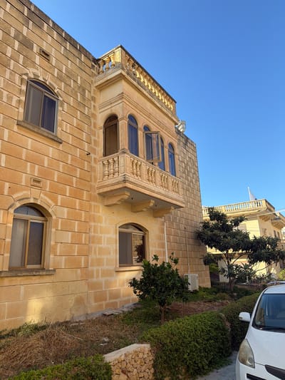 The image shows a two-story building constructed with light brown stone blocks. The building features several arched windows, with one on the first floor and two on the second floor. A prominent balcony with decorative balustrades is present on the second floor, with two of its windows open. Adjacent to this building, another building with a similar architectural style is visible, topped with a balustrade. The foreground includes a small garden with green bushes, dry grass, a small tree, and a white car parked on the right side. A stone wall is also visible in the lower part of the image. The scene is set under a clear blue sky, suggesting it is daytime. The overall impression is that of a residential area in a Mediterranean climate, likely in Xagħra, Malta, given the characteristic stone architecture.
