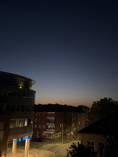 The image depicts an urban scene at dusk in Wilhelmshaven, Germany. The sky transitions from a deep blue to a lighter gradient, suggesting the setting sun. Dominating the foreground is a modern building with a curved, glass facade. Blue neon lights spell out "GESUNDHEITSZENTRUM" on this building, indicating it is a health center. Adjacent to this is a darker, older brick building with numerous windows, some of which are illuminated. A street runs between these buildings, with a crosswalk clearly visible. A dark-colored car is parked on the street. Trees and foliage are visible, partially obscuring some of the buildings. The overall impression is one of a quiet evening in a German city.