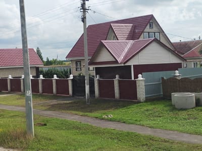 The image depicts a suburban street scene in Kargal, Russia, on an overcast day. In the foreground, a paved walkway runs alongside a grassy area with some weeds and a discarded concrete ring. A metal utility pole stands near the walkway, its surface partially obscured by stickers.

Dominating the midground are several houses with maroon metal roofs. The house directly behind the utility pole features a complex roofline with multiple gables and dormer windows. Its exterior walls are a combination of light-colored stone and beige vinyl siding. A dark red garage door is visible beneath one of the gables. This house is enclosed by a fence made of alternating maroon metal panels and white brick pillars topped with decorative white spheres. A black metal gate is set into the fence.

To the left of this house, another structure with a matching maroon metal roof is visible. Its exterior appears to be constructed from light-colored brick. To the right, a section of wooden fence with a scalloped top separates the property from another house, partially visible, with a similarly colored roof. In the far right of the midground, a few concrete rings are stacked on the grass.

The background shows more houses with red roofs, green trees, and a distant hilly landscape under a cloudy sky. Power lines crisscross the sky, supported by utility poles. The overall impression is one of a residential area undergoing development or maintenance.