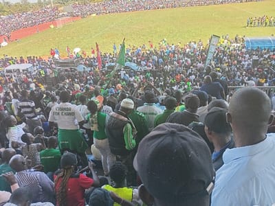 The image depicts a large, vibrant crowd gathered in an outdoor stadium under bright daylight, likely in Siaya, Kenya. The vast number of people fill both the stands and the field level, extending into the distance. Many attendees are dressed in green and white clothing, including shirts, scarves, and hats, and several green flags are prominently waved, suggesting a common affiliation or group identity. A red-colored running track encircles a green grass field, typical of a sports arena, with a blue dugout or shelter visible near the field's edge.
In the mid-ground, a person wearing a white t-shirt clearly displays the text "CHANDA TOK CHANO" on their back. Other visible objects include speaker systems or sound equipment mounted on a platform amidst the crowd, and a green banner with graphics, though its text is not legible. The weather appears sunny, contributing to the clear and bright illumination of the scene. The overall impression is one of a lively and well-attended public event, potentially a political rally, sporting event, or other large community gathering, characterized by a strong sense of collective enthusiasm and solidarity.