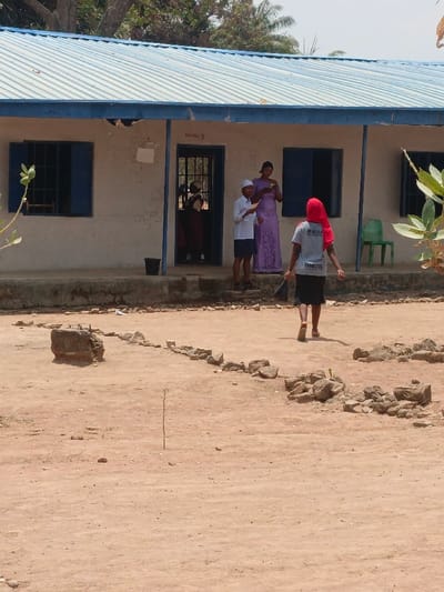 The image displays an outdoor scene in Mada, Nigeria, centered on a single-story building identified by a sign as "READY HALL B." The building features a light-colored facade, a blue corrugated metal roof, and a raised concrete porch. Two windows with open blue shutters and a dark, gridded metal door are visible on the front. A black bucket rests on the porch near the door, and a green plastic chair is positioned further right on the porch.

The foreground and midground consist of unpaved, light brown earth with scattered rocks forming an irregular boundary or outline. A thin, upright wooden stick is planted in the dirt in the immediate foreground.

Five individuals are present. In the foreground, a young female, clad in a grey t-shirt, red headscarf, and dark skirt, walks away from the viewer towards the building, holding a dark stick or broom. On the building's porch, a young male wearing a white cap, white long-sleeved shirt, and dark shorts stands, looking down at a white paper he holds. Adjacent to him, an adult female in a long, light purple patterned dress stands, examining a small object in her hands. Partially visible through the building's open doorway are two additional individuals, both wearing reddish-brown garments, positioned further inside the structure. The sky above the building is clear, with trees and foliage visible in the background.