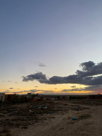 The image shows a sky filled with clouds, ranging from light to dark, against a gradient sunset. The ground is barren and littered with trash. Buildings are visible in the middle, creating a distinct line between the sky and the ground. The photo is rotated.