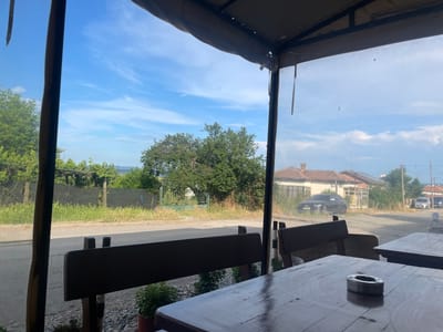 The image displays a daytime view from an outdoor seating area, likely a cafe or restaurant, located in Konstantinovo, Bulgaria. In the foreground, dark wooden tables and benches are visible. A silver-toned metal ashtray rests on the nearest table, reflecting ambient light. Small potted plants are situated on the ground beneath the benches. The view extends through a transparent plastic curtain or screen that serves as an outer wall for the seating area.
Outside, a paved road runs horizontally across the midground. To the left of the road, a grassy verge is present, bordered by a black mesh fence and further left by a green metal gate. Abundant green foliage, including trees and bushes, lines the road and fills the landscape. In the background, several houses with reddish-brown tiled roofs are discernible. A dark-colored SUV is parked or positioned near one house on the left side of the road, while two lighter-colored vehicles are visible further down the street on the right. The sky overhead is clear blue with sparse, elongated white clouds. No individuals are present in the scene.