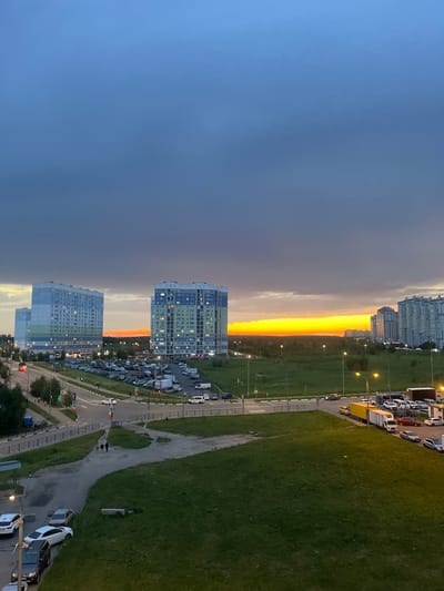 The image depicts a residential district in Domodedovo, Russia, during a dusky sunset. Dominating the skyline are several multi-story apartment buildings, one of which is light blue and white with green accents, and another similar in design but with blue and green sections. A third, taller building with a lighter facade is visible to the left. The sky above is a gradient of deep blue to dark grey, with a bright band of orange and yellow illuminating the horizon, suggesting the sun is setting.
Below the buildings, a moderately busy road curves through the scene, with a considerable number of parked cars filling a large lot. Some vehicles are in motion, including a red car, a white car, and a yellow truck with a white trailer. Streetlights are illuminated, casting pools of yellow light on the asphalt and surrounding grass. A patch of green grass slopes down from the foreground, with a paved pathway cutting through it, where two figures are seen walking with a dog. The overall environment appears to be a developed urban area with interspersed green spaces.