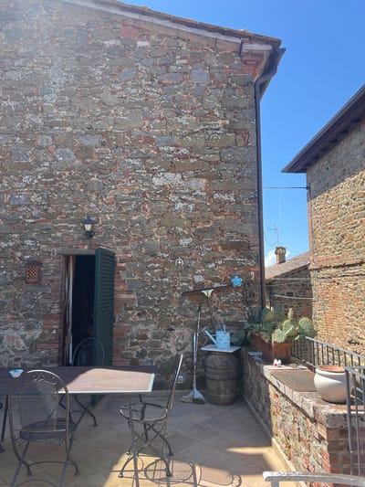 This image captures an outdoor scene in Sinalunga, Italy, centered on the exterior of a stone building under a clear blue sky. The building's facade is constructed with irregularly shaped stones, exhibiting a rough texture and varied coloration of grays and reds. An awning-covered window is visible on the wall, positioned to the side of a light fixture. Various decorative items, including a flower-shaped wall ornament and gardening tools, are affixed to the stone wall. A tiled patio area features a table and metal chairs, bathed in sunlight. Above, a brick structure is partially visible. Adjacent to the patio, a cactus plant is potted near a large barrel.