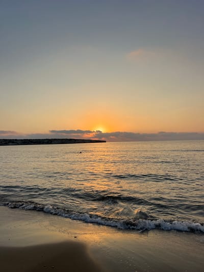 The image depicts a serene sunset over the sea in Ineia, Cyprus. The sun, partially obscured by a line of clouds, casts a warm golden glow across the sky and reflects on the calm water. A gentle wave washes onto a sandy beach in the foreground, creating a frothy white line. In the middle distance, a person is visible swimming in the sea. A rocky, elevated coastline stretches across the horizon on the left side of the frame. The overall scene conveys tranquility and the beauty of nature at dusk.