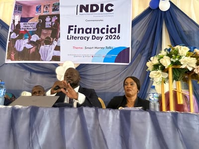 This image captures an indoor event, likely a seminar or conference on financial literacy, held in Unguwan Dosa, Nigeria. The main focus is a large banner in the background that prominently displays "NDIC Nigeria Deposit Insurance Corporation Protecting your bank deposits Commemorates Financial Literacy Day 2026 Theme: Smart Money Talks." An embedded picture on the banner shows a classroom scene, indicating educational activities. Below the event details, contact information including a toll-free help desk number, email, website, and social media handles like "@NDICNigeria" are visible.

In the foreground, two main figures are seated at a blue-draped table. A man, dressed in a suit, is speaking into a microphone, his hands gesturing expressively. To his right, a woman in a dark blazer appears attentive. Another man's head is partially visible behind a laptop to the speaker's left. Water bottles are placed on the table, and a decorative floral arrangement with white and blue flowers sits on the far right. The setting features blue and white drapes and balloons, contributing to a formal atmosphere for the financial education event.