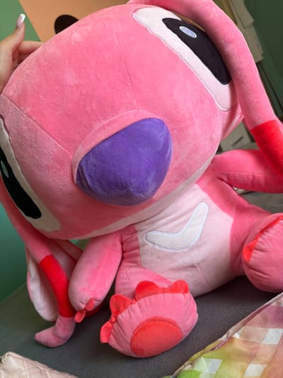 The image displays a large, pink plush toy, strongly resembling the Disney character Angel from Lilo & Stitch. The toy is predominantly a vibrant pink, with lighter pink sections on its face and stomach. Key features of the plush include large, oval black eyes with white highlights, a prominent purple nose, and reddish-pink accents on its inner ears, antennae tips, and paw pads. A distinctive white, boomerang-shaped patch with a lighter outline is visible on its stomach.
A human hand, characterized by neatly manicured nails with white tips, is seen gently supporting the plush toy near its head, suggesting interaction or placement of the item.
The setting appears to be indoors, likely a domestic space. Various textural elements compose the background: a dark gray textured fabric, possibly a sofa or bedding, is visible behind the toy. To the upper right, a colorful checkered fabric in shades of green, yellow, pink, and white, likely a blanket or pillow, adds a soft pattern. In the lower right, a cream-colored, quilted object, possibly a bag or cushion, is partially visible. The lower left background shows a smooth, greenish-blue surface, potentially a wall or floor.
The image captures a static scene of the plush toy, conveying a sense of comfort and a relaxed atmosphere. No specific activity is occurring, and there is no visible text within the frame. The location context provided for this media file is Sapareva Banya, Bulgaria.