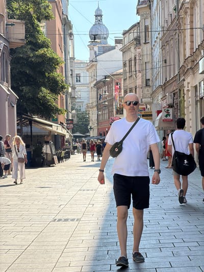 The image depicts a street scene in Sarajevo, Bosnia and Herzegovina, characterized by narrow, pedestrianized cobblestone pathways lined with historic buildings. In the foreground, a bald man wearing sunglasses, a white t-shirt, and black shorts walks towards the camera, carrying a green crossbody bag. Behind him, another man in a white t-shirt and grey shorts walks away, with a black messenger bag. Several other pedestrians are visible in the background, some walking, some standing near storefronts. The architecture features a mix of classical and more modern styles, with balconies, awnings, and signage indicating commercial activity. A prominent dome structure with a spire rises in the distance, a characteristic element of Sarajevo's skyline. Sunlight casts long shadows across the street, suggesting it is either early morning or late afternoon. The atmosphere appears to be one of a bustling urban environment on a clear day.