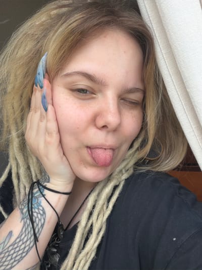 A blonde woman with dreadlocks and long, blue-painted nails is pictured in a close-up selfie. She is resting her left hand on her cheek and has her tongue sticking out, with her right eye winking. She is wearing a black t-shirt and has a tattoo on her left forearm. A light-colored curtain is visible to her right, and a dark background to her left. The setting appears to be indoors.
