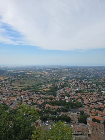 A high-angle, wide shot captures an aerial view of the City of San Marino, San Marino. The image shows a sprawling urban landscape characterized by terracotta-tiled roofs and dense clusters of buildings. Rolling hills, covered in a patchwork of green fields and agricultural plots, surround the city and extend into the distance, eventually meeting a hazy horizon. A winding road is visible within the urban area, with a few vehicles present. In the foreground, lush green trees partially obscure the view, suggesting the vantage point is from a higher elevation, possibly a hill or a nearby fortification. The sky above is a pale blue, with scattered white clouds. The overall impression is of a historic city nestled within a picturesque, hilly landscape.
