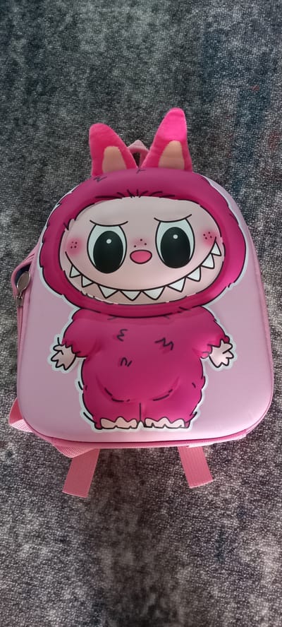 A pink backpack with a cartoon monster design is laid flat on a gray, textured surface, possibly a carpet. The backpack features a plush pink hood with floppy ears, and the front displays a large, smiling monster face with big black eyes, sharp white teeth, and rosy cheeks. The body of the monster is depicted in a darker shade of pink, with a fuzzy texture. The backpack has pink straps and a silver zipper. The background is a consistent gray texture, showing no distinct features or architectural elements that would indicate a specific location within Andzhero-Sudzhensk, Russia.