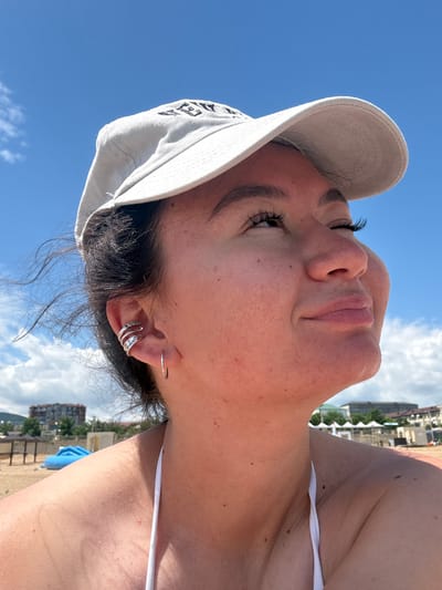 A medium shot, low-angle outdoor photo shows a young woman wearing a light-colored baseball cap and a white bikini top. She is looking up and to the right, with her eyes closed. Her right ear is visible, adorned with a silver ear cuff and a hoop earring. Her hair is dark and appears windblown. The background consists of a clear blue sky with scattered white clouds, and a beach scene with buildings and vegetation in the distance. The overall lighting suggests a bright, sunny day. The location is identified as Derbent, Russia.