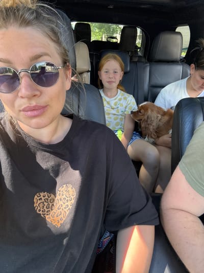 In Valday, Russia, a group of people and a dog are inside a car. The photo is taken from the perspective of the person in the front passenger seat, who is wearing sunglasses and a black t-shirt with a leopard print heart graphic. To their right, a young girl with red hair is seated in the back, wearing a yellow t-shirt with a lemon pattern. Next to her, a woman in a white t-shirt is holding a brown and white cocker spaniel. Another person, partially visible in the foreground on the right, is wearing a green t-shirt. The interior of the car appears to be black leather. Sunlight is casting a strong shadow and a rainbow-like light effect on the front passenger.