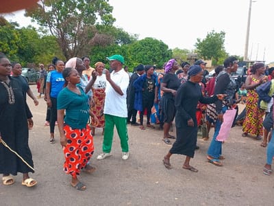A group of about 20 people, mostly women, stand in a street, likely in Ungwan Tanko, Nigeria. The street is paved and appears to be a main thoroughfare, with some trees and foliage in the background. Several people are talking and interacting, while some stand with arms crossed or hands on their hips. One man is wearing a green shirt and pants, and another woman is holding a pink bag. The group appears to be gathered for a meeting, event, or gathering.