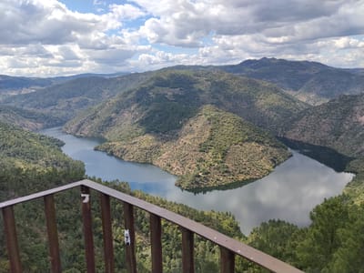 The image depicts a scenic view of the Douro River valley from a lookout point in Carrazeda de Ansiães, Portugal. A series of forested hills and mountains frame the winding river. The sky is partly cloudy, with patches of blue visible between the clouds. In the foreground, a metal railing of the viewing platform is visible, providing a sense of depth to the scene. The river reflects the sky, and the hillsides are covered in dense vegetation.