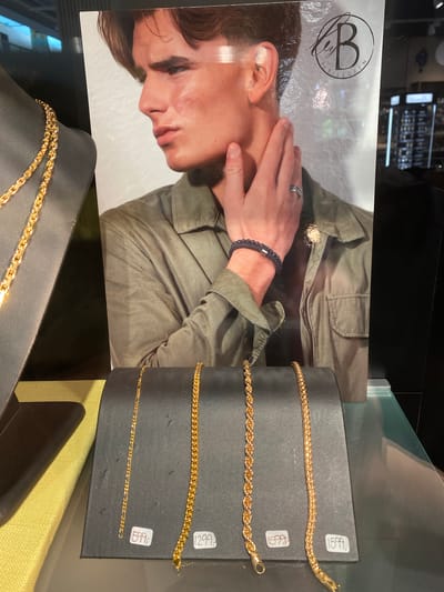 An advertisement for jewelry by Billgren is displayed. The advertisement features a close-up of a young man's face and shoulder, wearing a green jacket. He has a hand near his face, adorned with a ring and a beaded bracelet. A logo for "B by BILLGREN" is visible in the upper right corner of the advertisement. In front of the advertisement, on a black display stand, are several gold necklaces. Two necklaces are on display in the foreground, each with a price tag. The left necklace is a Figaro chain priced at 599. The middle necklace is a rope chain priced at 1299. The right necklace is a braided chain priced at 1599. Another gold necklace is displayed on a black mannequin bust to the left of the advertisement, but it has no visible price tag. The scene appears to be inside a jewelry store, possibly in Herning, Denmark, as indicated by the prices and the general presentation of retail merchandise. The overall lighting suggests an indoor retail environment.
