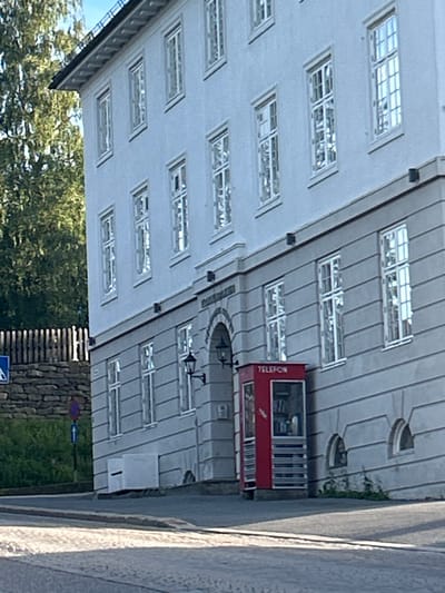 The image captures a portion of a light-colored, multi-story building in Kongsberg, Norway. The building has numerous windows with white frames, many reflecting the sky and surrounding trees. A distinctive bright red telephone booth, labeled "TELEFON," is situated at the base of the building. To the left of the building, there is a stone wall and a wooden fence, with greenery behind them. A blue traffic sign with an arrow is visible on a pole. The foreground shows a cobblestone street and a paved road, suggesting an urban or town setting. The lighting indicates it is daytime, with sunlight casting shadows.