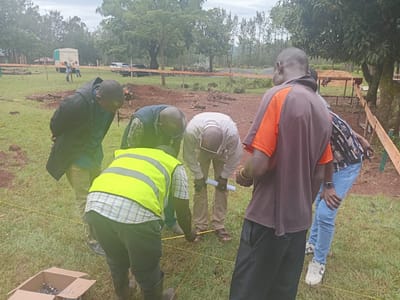 The image depicts a group of six individuals in an outdoor environment, likely the city of Idakho South ward, Kenya, based on the setting and individuals depicted. The group is gathered around an area marked with yellow string, possibly related to construction or surveying activities. One person is wearing a bright yellow safety vest. Several individuals are looking down at the marked area, possibly examining measurements or plans. One individual has a rolled paper in their hand. A box containing metallic nails or stakes is on the ground nearby. In the background, there is a white truck, some building materials, and more individuals standing around. The terrain appears to be grassy and slightly uneven. Some trees are visible in the background. The sky is overcast.