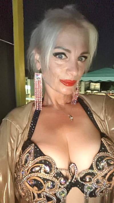 This is a medium shot selfie of a woman with short, platinum blonde hair, wearing bright red lipstick and heavy eye makeup with winged eyeliner. She has large, rectangular earrings with pink fringe that sparkle with rhinestones. She is also wearing a bejeweled black bra with gold accents and a shiny, gold satin-like jacket. A delicate silver necklace with a teardrop pendant is visible around her neck. Her expression is confident and direct, looking at the camera. The background is dark, suggesting it is nighttime, with a blurry yellow pole on the left and what appears to be a green tent or awning in the distance on the right, possibly indicating an outdoor event or venue. The image appears to have been taken in Kuşadası, Turkey, given the location context.