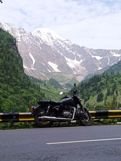 A dark-colored Royal Enfield motorcycle is parked on the shoulder of a paved road in Dhundi, India. The motorcycle is facing left, with its kickstand down. The road is empty except for the motorcycle and a yellow and black guardrail. Behind the guardrail, a steep, tree-covered mountainside rises, leading to snow-capped peaks in the distance under a cloudy sky. The environment is lush and green, indicating a mountainous region with some snow.
