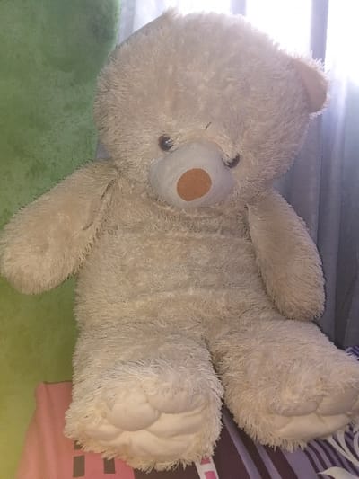 A tan-colored, plush teddy bear lies prone on a multicolored surface. The bear has a round body, small dark eyes, and a brown nose. Its fluffy fur is slightly disheveled. The surface underneath appears to be a mix of green fabric and a patterned material with pink, white, and dark stripes. The bear's limbs are outstretched, giving the impression it is relaxed.