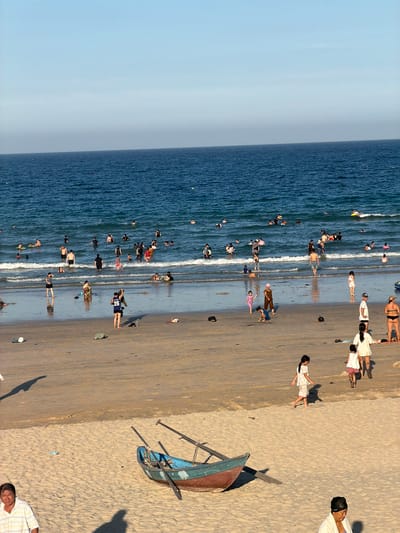 The image depicts a crowded beach scene in Cam Lam, Vietnam, likely during daylight hours given the bright sunlight and clear sky. The ocean is a deep blue, with moderate waves lapping at the shore. Numerous people are in the water, some swimming, others standing or wading. On the sandy beach, a variety of activities are taking place. Several individuals are walking or standing, while children are seen playing. A few boats, including a partially submerged blue and red one with oars, are present on the sand. Clothing, bags, and a plastic bottle are scattered across the beach. The overall atmosphere suggests a popular recreational area.