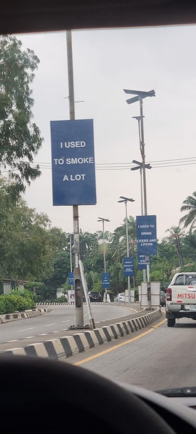 This is a street-level, eye-level, outdoor, daytime, medium shot taken from a vehicle, looking forward. The image captures a road in Ikoyi, Nigeria, with a prominent set of blue billboards on light poles. The billboards convey personal affirmations and past habits. The first billboard on the left reads "I USED TO SMOKE A LOT." Further down the road, another billboard states, "I USED TO DRINK LITERS AND LITERS." Additional, partially visible billboards suggest themes of change and aspiration, with phrases like "I REALLY WANTED TO MAKE A CHANGE" and "SO I BUILT MY DREAM." The road itself is paved and curves to the right, with clear lane markings and black-and-white striped traffic barriers. Lush green trees and palm trees line the sides of the road, indicating a tropical environment. A white Mitsubishi pickup truck is visible in the right lane, as are other vehicles in the background. The sky is overcast, suggesting a cloudy or hazy day. The overall scene depicts a public space in a Nigerian city adorned with motivational messaging.