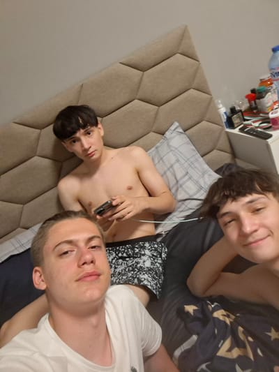 In Belashtitsa, Bulgaria, three young males are positioned on a bed, with a nightstand containing various items visible to the right. The male in the foreground is wearing a white t-shirt and looking towards the camera with a slight smile. To his left and slightly behind him, another male is shirtless, wearing patterned shorts, and holding a mobile phone. He is looking at the camera with a neutral expression. The third male is in the background, also shirtless and wearing patterned shorts, with his head resting on a pillow and looking to the side. The room appears to be a bedroom, with a large, upholstered headboard behind the individuals and a patterned duvet on the bed. The lighting suggests it is daytime.