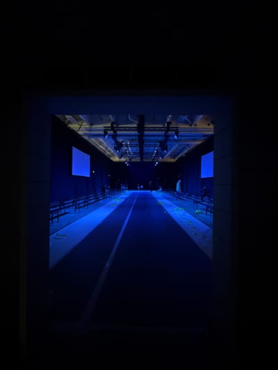 The image captures an indoor event venue, likely located in Issy-les-Moulineaux, France, characterized by a dominant blue lighting scheme. A long, dark, central runway or track, marked with a lighter longitudinal line, extends from the foreground toward the background. On both sides of this runway, multiple rows of dark benches are arranged as seating. The floor areas immediately adjacent to the runway and beneath the benches are bathed in blue light. Overhead, an extensive grid of industrial lighting fixtures and spotlights illuminates the space. Two large, rectangular screens, displaying a solid blue color, are mounted vertically on the side walls further into the venue. Several indistinct human figures are visible; some appear to be standing or moving near the seating areas, while at least one person is seated on the benches on the far right. Green, organic material is scattered across the blue-lit floor areas flanking the runway, particularly on the right side. Yellow arrow-shaped markings are present on the blue-lit floor segment to the left. The scene is framed by a dark, rectangular architectural opening in the immediate foreground.