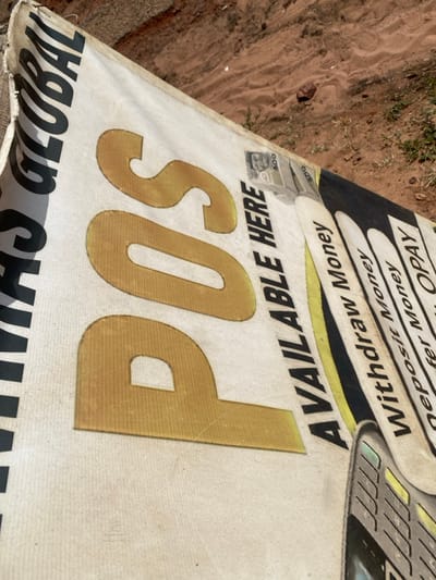 This image depicts a white, rectangular banner laid partially on reddish-brown dirt ground with sparse green vegetation, presumably in Okura, Nigeria. The banner displays large gold text "POS" centrally. To the left, vertically oriented black text reads "GLOBAL" (partially visible). To the right of "POS", black text states "AVAILABLE HERE". Further right, a vertical list in black text includes "Withdraw Money", "Deposit Money", "Transfer Money", and "OPAY" (partially visible). A graphic representation of a stack of Nigerian 500 Naira notes is visible above "AVAILABLE HERE", and the bottom portion of a mobile Point of Sale (POS) terminal is depicted at the lower right. The banner shows signs of wear and slight discoloration. No individuals are present in the scene.