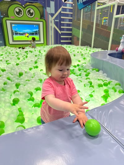 A young child in a pink shirt is in a ball pit filled with green and white balls, located inside an indoor playground. The child is reaching for a green ball resting on a blue padded surface. A screen with a cartoonish frog design shows children playing a soccer game. The playground includes netting and structures. The location of the scene is Перовское сельское поселение, Ukraine.