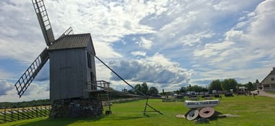The image depicts the Angla Windmill complex in Angla, Estonia. In the foreground, a large, weathered wooden windmill stands on a stone base. Its sails are extended, with one sail broken. A ladder leads up to a wooden balcony on the side of the windmill. In front of the windmill, a wooden fence runs diagonally across the frame. To the right of the windmill, a large wooden beam with text inscribed on it and several millstones are visible on the grass. The background shows a parking lot filled with cars and campers, a stone building with a red roof, and a line of trees. Several people are scattered throughout the scene, some walking near the vehicles and others near the stone building. The sky is filled with dramatic white and gray clouds.
