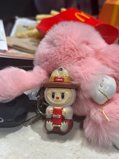 A fluffy pink bunny keychain with white ears and a smaller bunny figurine holding a Coca-Cola can are on a keyring. A car key is also attached to the keyring. In the background are McDonald's fries and other items. The surface underneath is light-colored.