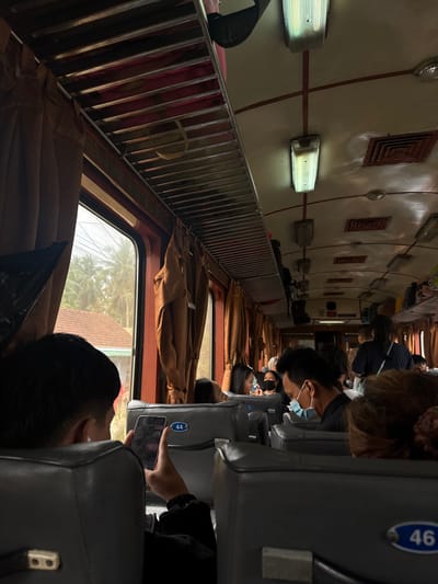 The image captures the interior of a train compartment, likely in Vietnam, given the presence of palm trees and a tropical atmosphere visible through the window. The train car is filled with passengers seated in rows on both sides. The seats are upholstered in a dark material, and overhead luggage racks are visible. Lighting fixtures are mounted on the ceiling, casting a warm glow on the scene.
In the foreground, a person wearing a black jacket is looking at their phone, which is held in their right hand. The phone screen is illuminated, showing some text or graphics. The person's head is turned slightly away from the camera, revealing the back of their head and the side of their face.
Further down the aisle, other passengers are visible, some wearing face masks. The overall impression is that of a crowded but orderly train journey, with people engaged in their own activities or interacting with each other. The exterior view through the window suggests a rural or semi-urban setting with lush vegetation.
