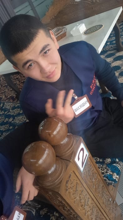 The image captures a young male seated indoors, likely within a cafe or restaurant setting, in Chinabad, Uzbekistan. The subject, with short dark hair, wears a dark blue vest bearing a name tag that reads "ШАХЗОДБЕК" (Shakhzodbek) in Cyrillic, over a dark long-sleeved shirt and dark trousers. He exhibits a faint smile, with his right hand partially raised and fingers splayed. In the foreground, positioned to his left, is a decorative wooden stand with intricate carvings, topped by two glossy brown wooden spheres. A white square label with the red numeral "2" is affixed to the side of this stand. The floor is covered by a patterned rug or carpet in shades of blue and white. In the background, a white rectangular table supports a wicker basket, a white tissue box, and a round, patterned coaster. An ornate wooden chair is partially visible behind the table.