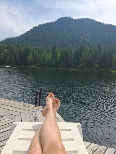 A person's legs and feet are shown from the knees down, resting on a white lounge chair on a wooden dock. The legs are crossed at the ankles and appear wet. The dock extends to the edge of a dark, rippling lake. In the background, a dense forest covers a large mountain under a partly cloudy sky. A small white paddleboat with two people can be seen on the lake in the distance. The scene is located in Vydrino, Russia.