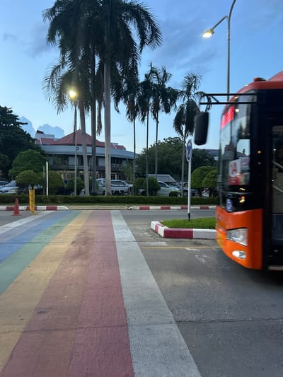 The image depicts a street scene in Tha Khlong Town Municipality, Thailand, at dusk. The foreground features a rainbow-colored crosswalk, with stripes of purple, blue, green, yellow, and pink. To the right, an orange bus is in motion, its front visible, with its headlights on. The bus has a black interior and an orange-tinted windshield. A blurry black object hangs from the bus's windshield. A streetlamp with an illuminated bulb stands behind the bus. Palm trees with lush green foliage are scattered throughout the scene, some reaching towards the twilight sky. In the background, a building with a red roof and light-colored walls, featuring several windows and vertical columns, is visible. Several parked cars and a white pickup truck are also present. The environment suggests an urban setting with vegetation.