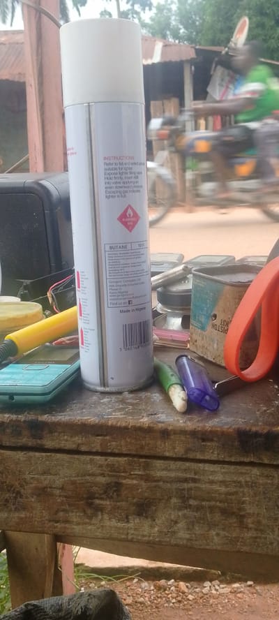 A butane gas refill can, labeled "Gongoni Company Limited," stands upright on a weathered wooden surface. The can is white with text in various colors and features a flammable gas warning symbol. Next to the can are two lighters, one green and one purple, and an orange scissor handle. A light blue mobile phone is visible to the left of the can. In the background, out of focus, a person on a motorcycle is visible, along with a corrugated iron roof structure, suggesting an outdoor or semi-outdoor setting in Baissa, Nigeria. The ground appears to be dirt and small stones. The lighting suggests it is daytime.