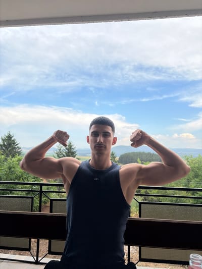 A young man with short dark hair and a mustache stands on a balcony, flexing his biceps. He is wearing a dark blue sleeveless shirt and dark pants. The background shows a lush green landscape with rolling hills and a blue sky with scattered clouds. The balcony has a dark railing and a dark wooden surface. The location is identified as Vladislavtsi, Bulgaria.
