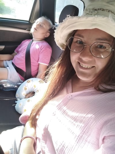 The image shows two people in the back seat of a car. On the left, a young girl is asleep, wearing a pink t-shirt and gray shorts. She is secured by a black seatbelt. Next to her, a blue travel pillow with a bee and daisy pattern lies on the seat. A clear plastic water bottle is also visible.
On the right, a woman is taking a selfie. She is wearing glasses and a light-colored bucket hat. Her top is a light pink, textured button-down shirt. The woman appears to be smiling. Her hair is long and reddish-brown.
The scene appears to be set during the daytime, with sunlight filtering into the car, creating bright highlights. The overall mood seems relaxed and casual, possibly during a road trip. The location is identified as Grabovo, Russia, but no specific landmarks are visible in the image. There is some faint text visible on the inside of the woman's hat, but it is partially obscured and difficult to read.