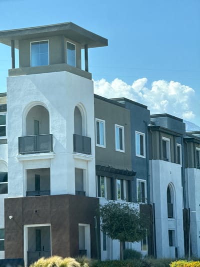 This image shows a multi-story building with a modern architectural style. The building features a mix of stucco finishes in white, brown, grey, and blue. There are multiple balconies with glass railings, arched windows, and rectangular windows. A small, leafy tree is visible in front of a section of the building. The sky is blue with scattered white clouds, and the background suggests a mountainous or hilly terrain. The image appears to be taken from a slightly low angle, looking up at the building. The location is identified as Fontana, United States of America.