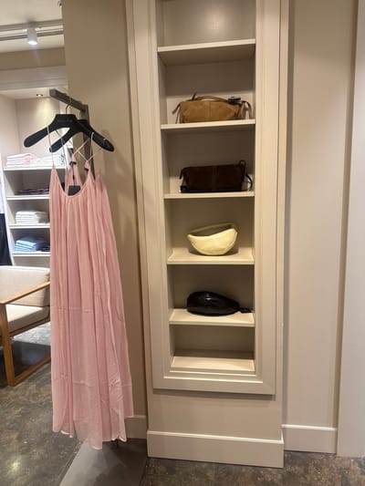 The image shows a built-in closet with four compartments in the upper section, holding handbags. The first compartment contains a tan suede handbag, the second a dark brown suede handbag, the third an off-white leather handbag, and the fourth a black leather handbag. Below the closet's upper section, clothes hang on a rack, including a long pink dress. The clothes hanging are partially visible within the closet's interior. The floor is grey patterned tile. The closet appears to be inside a residence or retail space, but the location in Leiria, Portugal, cannot be determined from this image alone.
