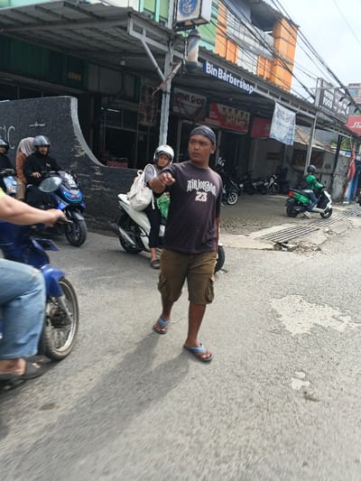 This image captures a daytime street scene in Margahayu, Indonesia. In the foreground, a male wearing a dark baseball cap, a dark t-shirt with "AIR JORDAN 23" text, brown cargo shorts, and blue flip-flops walks towards the viewer's right, extending his right arm forward in a pointing gesture. To his left, the lower torso in blue jeans and a portion of a blue motorcycle are visible. In the mid-ground, a dark blue scooter is stopped or moving slowly, carrying a male rider in a dark helmet and a female pillion passenger in a light brown hijab. Adjacent to them, a white scooter is visible with a female rider wearing a light helmet and a light-colored backpack. Further in the background on the right, a person in a green helmet and green jacket operates a green scooter, consistent with a delivery service. The background features a row of commercial buildings, including "Bin Barbershop" identifiable by its sign and a traditional barber pole, and "WIMI SHOP." Other visible signage includes "PROTEIN FOOD" and "J&T." The street is paved with asphalt, showing some wear and a storm drain grating on the right side. Numerous overhead utility wires are present across the scene.