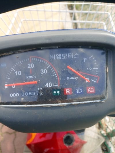 A close-up, high-angle view captures a dusty black instrument cluster belonging to an electric scooter or similar compact vehicle, located in 강화군, South Korea. The speedometer's red needle indicates approximately 10 km/h, with the dial marked up to 40 km/h. A digital odometer displays "005376 km". An analog battery gauge shows a red needle positioned towards "E" (Empty), indicating a low battery charge. The cluster prominently features the Korean text "비엠모터스" (Biem Motors) at the top. Other visible indicators include "Ready" on the left, unilluminated green left and right turn arrows, an unilluminated red "R" for reverse, an unilluminated blue headlight symbol, and a red battery warning symbol. A portion of a white wire mesh basket is visible above the cluster, while a red section of the vehicle's body, incorporating a black circular component (possibly a charging port or keyhole), is visible below. The underlying surface appears to be reddish-brown paving stones.