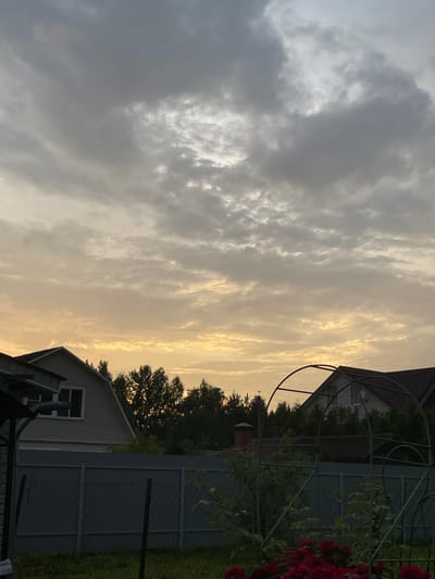 The image depicts an outdoor scene in a rural setting, likely a dacha or garden in Russia. The composition is dominated by the sky, which is filled with dramatic clouds. On the left, the clouds are a mixture of dark grey and bright white, suggesting a dynamic weather system. Towards the right, the sky transitions to a warm, golden hue, indicating either sunset or sunrise.

In the foreground and midground, elements of a residential area are visible. There are parts of buildings, including roofs and siding, suggesting houses or sheds. A grey fence encloses a portion of the property, and some greenery, including trees and a cluster of red roses, are present. A metal garden structure, possibly an arbor or trellis, is also visible, draped with ivy or another climbing plant.

The time of day appears to be late afternoon or early evening, given the warm, low-angled light illuminating the clouds. The weather seems to be changing, with the presence of both heavy clouds and bright sunlight breaking through. There are no people or animals visible in the image. The overall mood is tranquil and somewhat dramatic, owing to the striking sky. No text is discernible in the image.