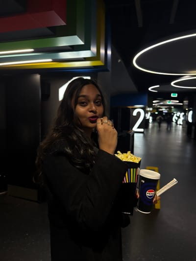 In a low-lit theater, a woman with dark hair and bold lipstick poses with popcorn and a Pepsi. Rings adorn her hand as she looks towards the viewer. Behind her, abstract, colorful wall decor adds visual interest. The theater's hallway is visible, marked by illuminated, curved light fixtures and seat numbers. People are faintly seen in the background, suggesting a lively atmosphere.