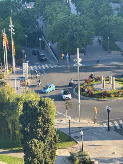 This is an elevated view of a roundabout and surrounding streets in Tarragona, Spain.
**Environment:** The scene is bathed in daylight. Lush green trees dominate the landscape, framing the urban area. The sky is obscured.
**Objects:**
* **Roads:** A roundabout occupies the center of the frame. Pedestrian crossings are clearly marked on the roads leading into the roundabout.
* **Vehicles:** Several vehicles are present including a blue car, a white car, and two dark-colored cars on the uphill road to the left.
* **Motorbikes:** There are three visible motorbikes, scattered around the roundabout.
* **Street Furniture:** Lamp posts and street signs are placed along the roads and pavements. A small concrete structure with vegetation sits on the edge of the roundabout.
* **Flags:** On the left, three flags are on flagpoles.
* **Buildings:** A building is seen on the upper right side.
**People:**
* Pedestrians are scattered in the upper half of the frame, and atop the hill, walking and sitting on benches.
* People are riding motorbikes in the roundabout.
**Location Details:** The layout of the roundabout and the surrounding buildings/landscape match that of the roundabout near the Balcó del Mediterrani in Tarragona, Spain. The flags likely belong to the province or the community of Catalonia.