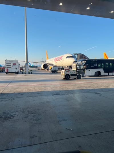 In Pendik, Turkey, at an airport tarmac, a Pegasus Airlines airplane is visible. The aircraft, bearing the registration "pegasus.com" on its fuselage, is positioned next to a walkway and a COBUS airport bus. In the foreground, a small, white ground support vehicle, identified as "TCE" and labeled "GROUND SERVICES," is present. The scene is illuminated by clear daylight, with a bright blue sky overhead.
